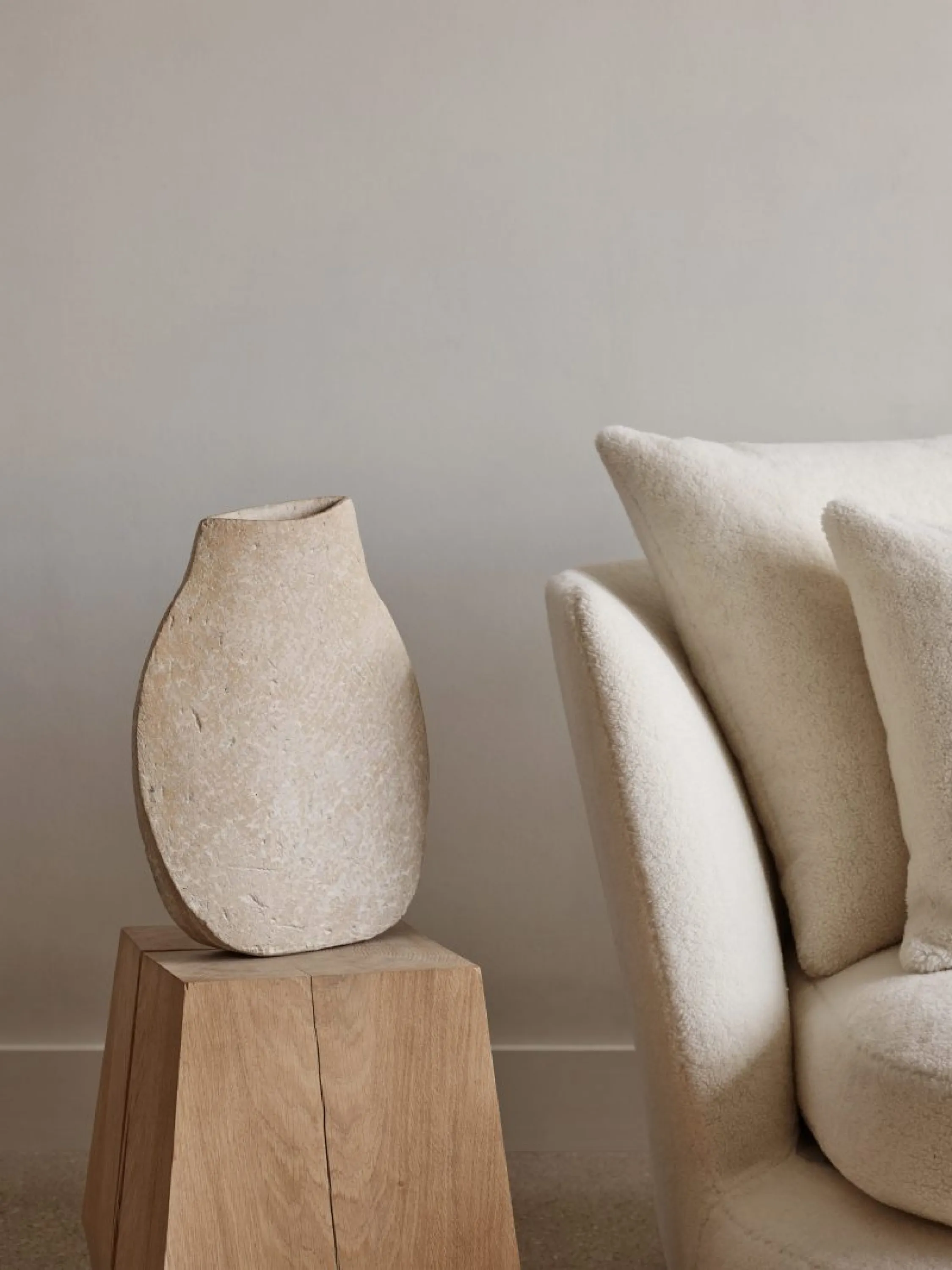 A tonal juxtaposition of textures: a rough stone vase, a wooden stool, and a soft armchair.